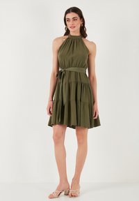 Abito senza maniche di colore verde oliva con colletto all'americana, vita stretta e gonna a strati. Tessuto morbido con un dettaglio annodato in vita.
