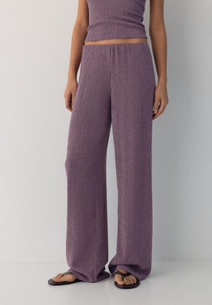 Frau trägt hochtaillierte, weitbeinige, strukturierte Lavendelhosen mit passendem cropped ärmellosem Oberteil und schwarzen Flip-Flops.