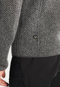 Szary sweter o teksturowanej strukturze, dopasowany do sylwetki, z małą czarną metką na dole. Stylizowany z czarnymi spodniami, co podkreśla kontrast.