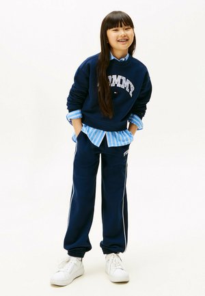 Młoda dziewczyna z długimi ciemnymi włosami, ubrana w granatowy sweatshirt od Tommy, dopasowane spodnie dresowe, paskowaną niebieską bluzkę i białe sneakersy, uśmiechająca się z rękami w kieszeniach.