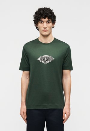 Άνδρας με σγουρά μαλλιά που φοράει μια σκουροπράσινη κοντομάνικη μπλούζα με λευκό λογότυπο "Emporio Armani Milano 1981", στέκεται μπροστά σε απλό φόντο.