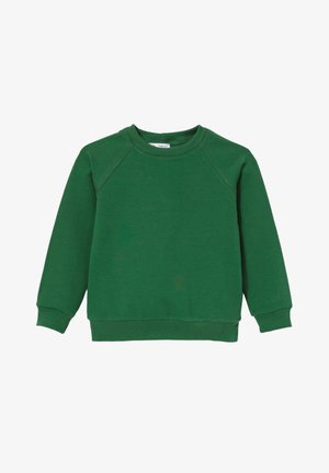 Uśmiechający się młody chłopiec w zielonej bluzie, niebieskich dżinsach i białych trampkach stoi z rękami w kieszeniach na jednolitym tle.