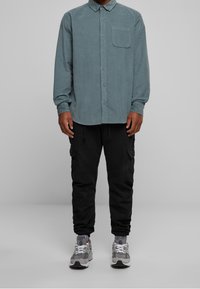 Langarmige, knoopsluiting shirt in gedempt teal met een borstzak, gecombineerd met zwarte cargobroeken en grijze sneakers met witte veters.