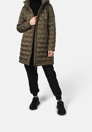 Personne portant une longue doudoune matelassée vert olive, un pantalon noir et des baskets noires, debout devant un fond blanc.