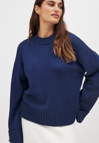 Navy blue knitted sweater with a crew neck, ribbed cuffs, and hem. Features a relaxed fit and a soft texture, paired with a white textured skirt.