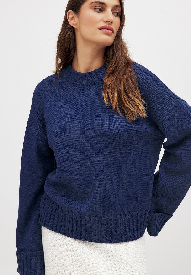Navy blue knitted sweater with a crew neck, ribbed cuffs, and hem. Features a relaxed fit and a soft texture, paired with a white textured skirt.