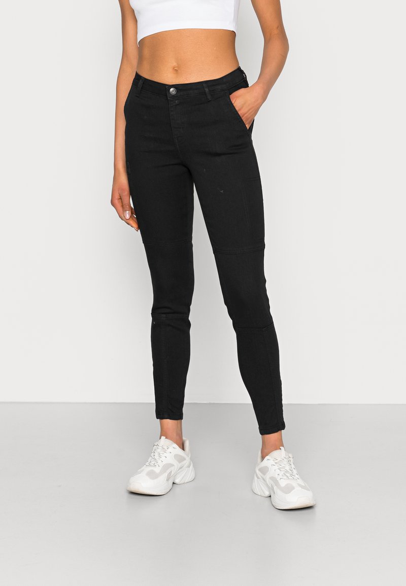 Calças skinny pretas em denim, com uma cintura de altura média, bolsos frontais e um acabamento liso, acompanhadas de sapatilhas brancas.