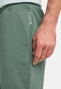 Grüner Sweatshirtstoff, Seitentasche mit Nähdetail und einem hellblauen Etikett am Bund. Hand ruht an der Seite.