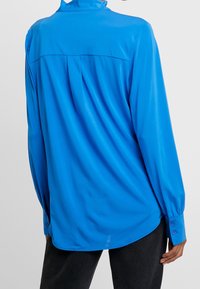 Blå långärmad skjorta med högre krage, tillverkad av glänsande tyg. Har knäppta manschetter och en ryggkil för extra detalj.