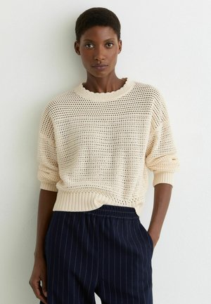 Kobieta z krótkimi włosami, ubrana w kremowy sweter z dzianiny i granatowe spodnie w prążki, stoi na tle białej ściany, jedną ręką wsuniętą w kieszeń.