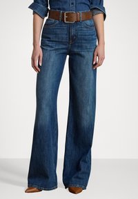 Vide benbukser i denim, med et brunt bælte med en firkantet metalsp�ænde. Bukserne har let falming og en lige skæring ved kanten.