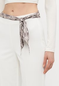 Sciarpa con motivo a pelle di serpente legata in vita, caratterizzata da sfumature di grigio e crema, abbinata a pantaloni bianchi a vita alta. Tessuto morbido.