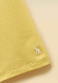 Coin de tissu jaune avec une fine couture et un petit lapin blanc brodé près du bord sur un fond beige clair.