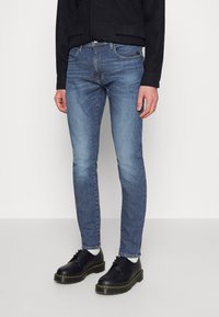 Jeans azules desgastados con un ajuste slim, diseño de cinco bolsillos y un ligero desgaste. Combinados con zapatos de cuero negros y calcetines blancos.