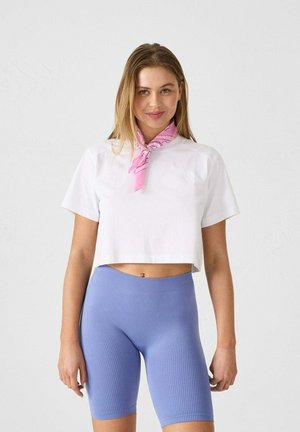 Camiseta blanca corta con mangas cortas, combinada con pantalones cortos de ciclismo de canalé en azul claro. El conjunto presenta un pañuelo rosa alrededor del cuello.