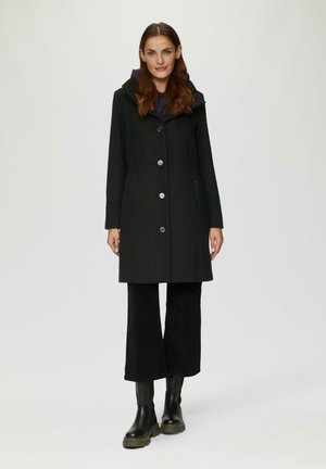 Black coat with a hood, featuring a button front and two side pockets. Worn over dark flared pants and black chunky-sole boots.