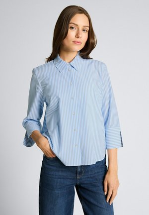 Femme portant une chemise à rayures bleu clair et blanc à boutons, manches trois-quarts, et un jean bleu foncé, debout avec une main dans la poche.