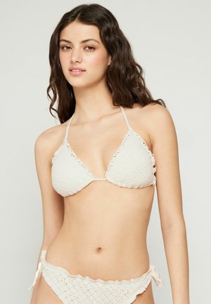 Jeune femme aux longs cheveux bruns ondulés portant un bikini blanc texturé avec des liens sur les côtés, debout devant un fond clair uni.