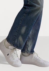 White leather sneakers with a smooth texture, featuring perforated detailing and a black logo on the side, worn with blue denim jeans.