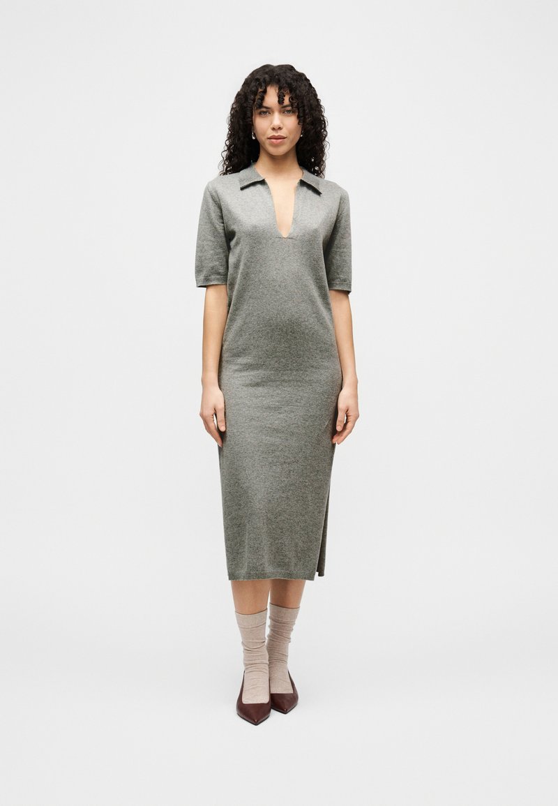 Γυναίκα με σγουρά μαλλιά, που φοράει ένα γκρι πλεκτό μίντι φόρεμα με βαθύ V-λαιμόκοψη, μανίκια μέσης μήκους, μπεζ κάλτσες και καφέ μυτερά παπούτσια, στέκεται μπροστά σε λευκό φόντο.