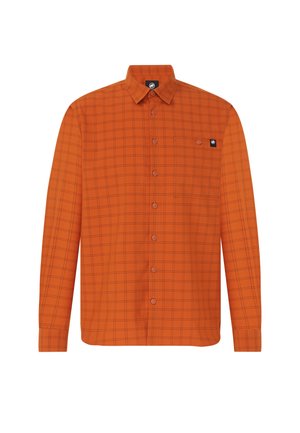 Mand med mørkt hår bundet tilbage iført en orange ternet skjorte over en hvid top, brune bukser og sorte sko, der står mod en ensfarvet baggrund.