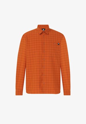Mand med mørkt hår bundet tilbage iført en orange ternet skjorte over en hvid top, brune bukser og sorte sko, der står mod en ensfarvet baggrund.