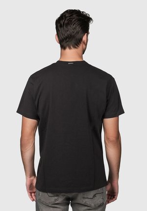 Hombre de cabello oscuro, vestido con una camiseta negra de manga corta sencilla y vaqueros grises, de pie de espaldas contra un fondo claro y uniforme.