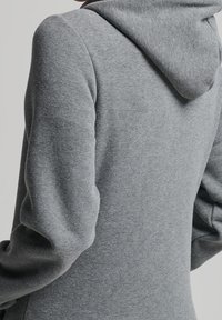 Persona che indossa una felpa grigia semplice vista di spalle con il cappuccio abbassato, evidenziando la texture del tessuto e i dettagli delle cuciture.
