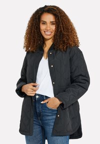 Chaqueta negra acolchada con cuello redondo, botones a presión y mangas largas; presenta un patrón texturizado y se lleva sobre una camiseta blanca.