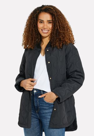 Zwarte gewatteerde jas met een ronde hals, drukknoopsluiting en lange mouwen; heeft een geperforeerd patroon en wordt gedragen over een wit shirt.