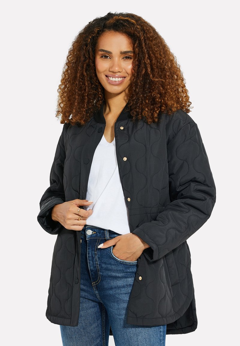 Chaqueta negra acolchada con cuello redondo, botones a presión y mangas largas; presenta un patrón texturizado y se lleva sobre una camiseta blanca.