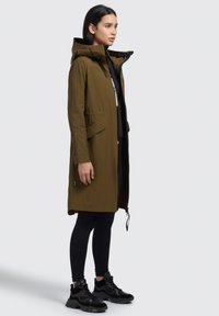Olivegrön lättviktsparka med huva, dragkedjestängning och sidofickor, ihopparad med svarta leggings och sneakers. Slät textur och modern design.