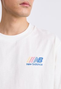 Tricou alb din bumbac cu logo-ul albastru "new balance" și dungi în galben, roz și albastru. Guler rotund și mâneci scurte.