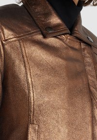 Bronzová metalická bunda s pevně strukturovaným límcem, výrazným prošíváním a patentkami, která vyzařuje hladký, texturovaný vzhled.