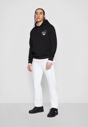Black hooded sweatshirt with front pocket and blue graphic, layered over a pink and white striped shirt. Paired with white pants and black sneakers.