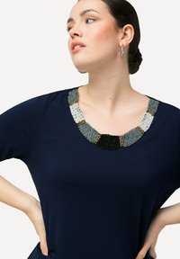 Top blu navy con scollatura rotonda caratterizzato da dettagli di perline intricate nei colori grigio, nero e metallici, texture morbida e maniche corte.