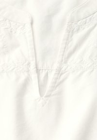 Top in cotone bianco con scollatura a V e dettagli ricamati lungo il collo e le cuciture delle spalle; texture liscia con una leggera lucentezza.