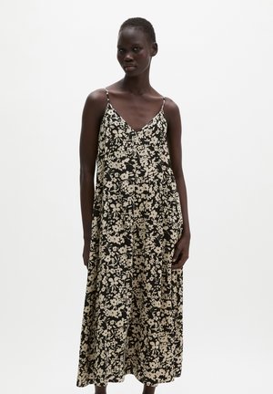 Femme portant une robe maxi fleurie noire et blanche avec des fines bretelles, debout devant un fond blanc uni.