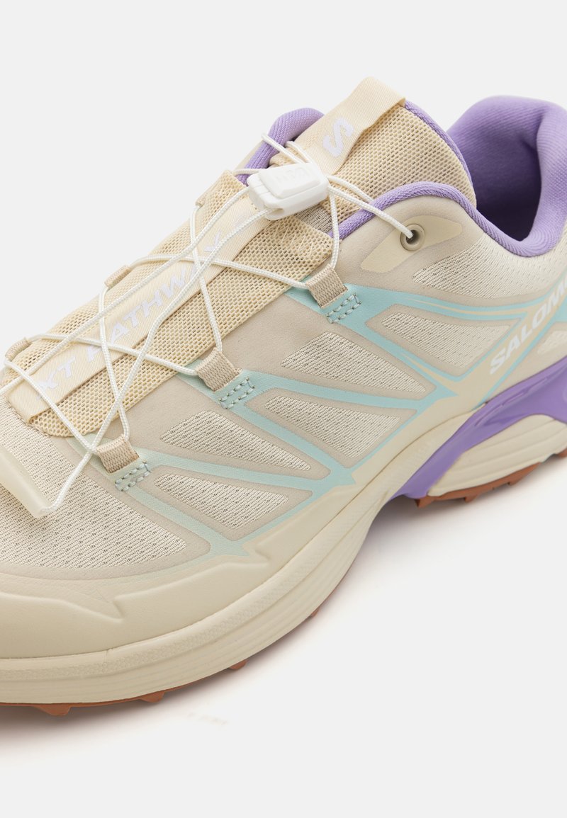 Zapatilla de running Salomon con una parte superior beige ligera, acentos de malla, detalles en azul claro y lavanda, y una suela de goma texturizada.