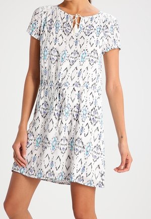 Biała sukienka z krótkim rękawem z niebiesko-czarnymi abstrakcyjnymi wzorami, z wiązaniem przy szyi i ściągaczem w talii, noszona przez stojącą modelkę.