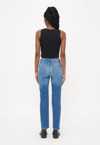 Svart ermeløs topp og lys blå rette jeans. Denimen har en jevn tekstur med baklommer. Personen har på seg svarte sko.