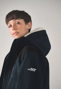 Tumesinist kapuutsiga jakk, millel on tõmbluku sulgumine, mustad aktsendid õlgadel ja valge "SUPER DRY" tekst varrukal, pehme sisemine vooder.