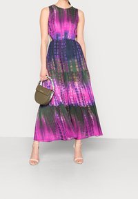 Robe longue avec un motif tie-dye vibrant violet, vert et rose, dotée d'un corsage ajusté et d'une jupe fluide, agrémentée d'un petit sac à main.