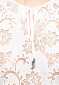 Λευκό ύφασμα με μπεζ φλοράλ δαντελένιο σχέδιο και δεμένη λευκή κορδέλα με ασημένια γούρια που κρέμονται στη λαιμόκοψη.