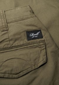 Primer plano del bolsillo de unos pantalones caqui con una etiqueta rectangular negra que dice "Reell est. 1997" cosida encima del bolsillo.