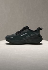 Μαύρο αθλητικό παπούτσι με χοντρή σόλα, μαύρα κορδόνια, επάνω μέρος από διχτυωτό υλικό με σχέδιο και τη λέξη «Palm Angels» στο πλάι, τοποθετημένο σε στρογγυλή φωτεινή επιφάνεια.