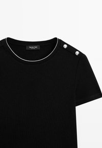 Chemise noire à manches courtes avec encolure ronde, passepoil blanc contrastant et deux boutons décoratifs argentés sur l'épaule droite.