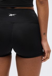 Γυναίκα που φοράει μαύρο αθλητικό σορτς Reebok και μαύρο crop top, με θέα στην πλάτη και το δεξί χέρι, όπου υπάρχει ένα μικρό τατουάζ με το γράμμα "M" στον καρπό.