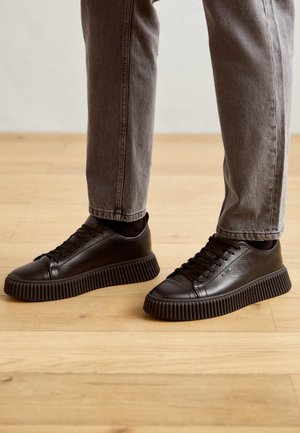 Person trägt schwarze, strukturierte Ledersneakers mit dicken, profilierten Sohlen und graue Hosen und steht auf einem Holzboden.