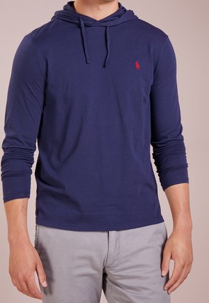 Man met een marineblauwe hoodie met lange mouwen en een rood logo op de borst, en lichtgrijze broek, staand tegen een effen achtergrond.